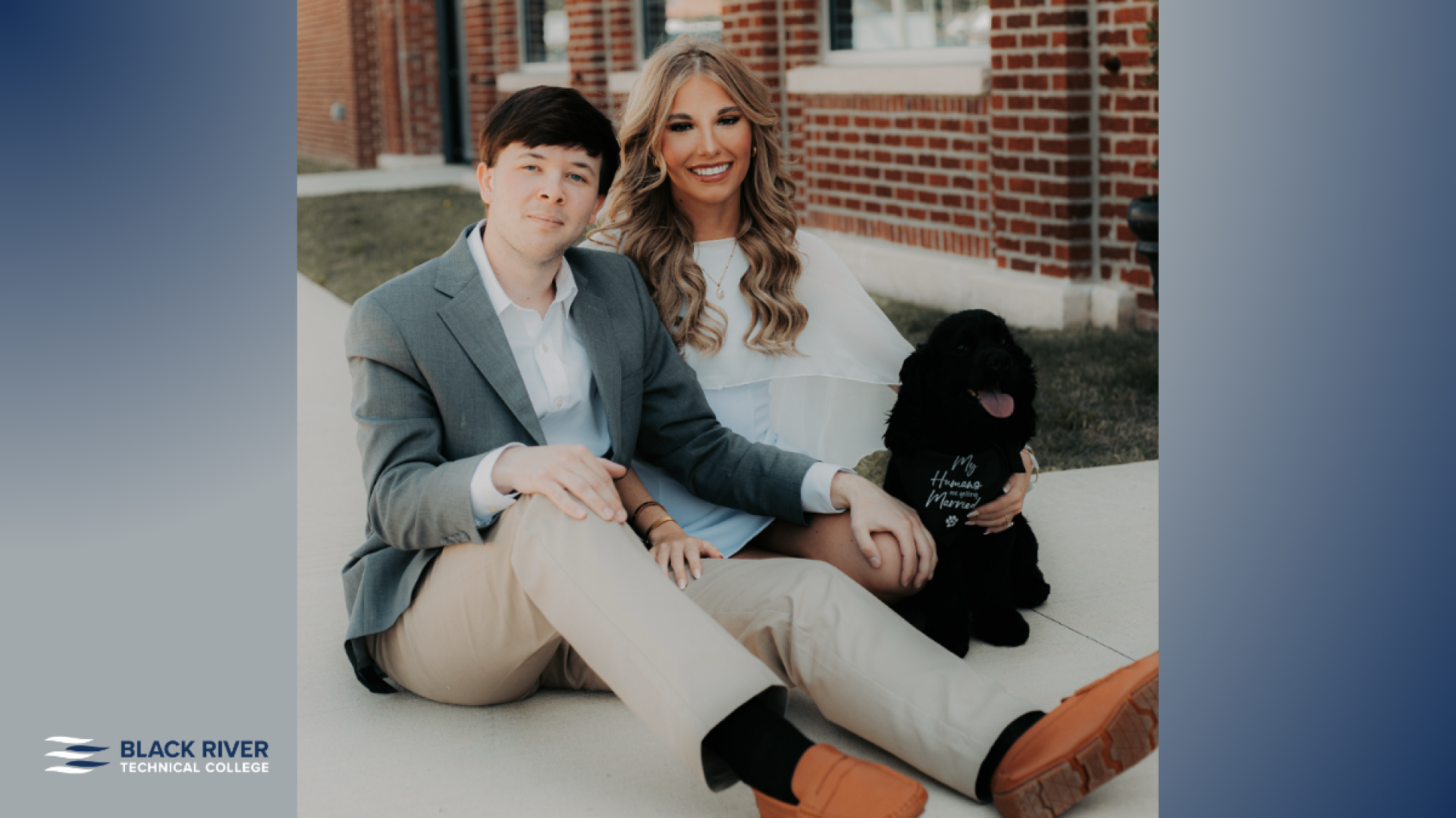 A young man in a gray blazer and a young woman in a white top sit together outdoors with a black dog wearing a bandana between them, in front of a brick building, with Black River Technical College branding.